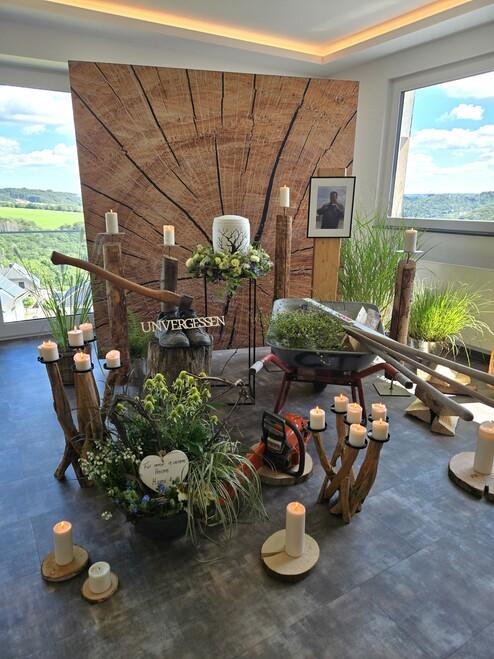 Blumengestecke und weiße Kerzen vor Holztafel neben Schubkarre für Bestattung