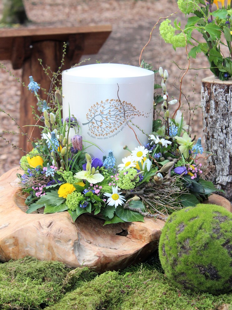 Wei&szlig;er Beh&auml;lter mit floralem Dekor, umgeben von bunten Fr&uuml;hlingsblumen auf einem Holzst&uuml;ck im Wald.